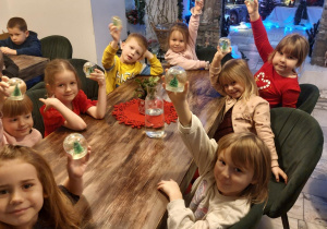 Grupa dzieci siedzi przy stoliku i podnosi do góry wykonane przez siebie na warsztatach plastycznych "kule śnieżne". Ujęcie 1