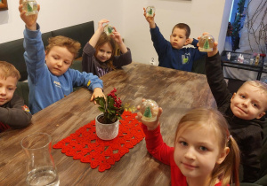 Grupa dzieci siedzi przy stoliku i podnosi do góry wykonane przez siebie na warsztatach plastycznych "kule śnieżne". Ujęcie 2