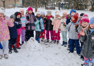 Grupa dzieci pozuje do wspólnego zdjęcia w zimowej scenerii ogrodu przedszkolnego.