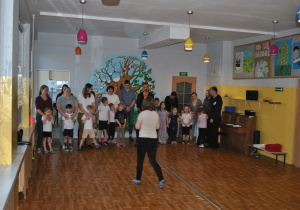 Rodzice i dzieci stoją razem w końcu sali gimnastycznej. Nauczycielka wyjaśnia zadanie.