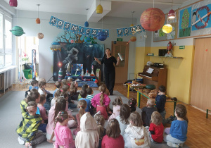 Grupa dzieci siedzi na dywanie i przygląda się nauczycielce opowiadającej o muzyce.