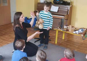 Chłopiec i nauczycielka prezentują grupie dzieci ciekawy instrument muzyczny.