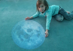 Dziecko siedzi na dywanie i obejmuje wyświetloną sylwetkę księżyca.