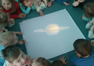 Dzieci siedzą na dywanie i przyglądają się wyświetlanej projekcji planety.