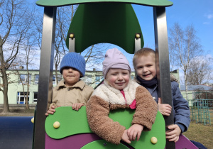 Dzieci z grupy 1 bawią się w ogrodzie przedszkolnym wczesną wiosną. Ujęcie 3. Troje dzieci uśmiecha się do zdjęcia stojąc na ogrodowej zabawce.