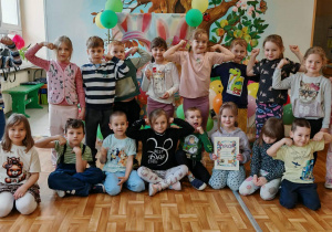 Grupa dzieci pozuje w przedszkolu do wspólnego zdjęcia z dyplomem i pucharem.