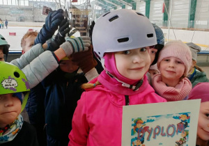 Grupa dzieci już na lodowisku cieszy się z dyplomu. Dziecko w różowej kurtce pokazuje dyplom