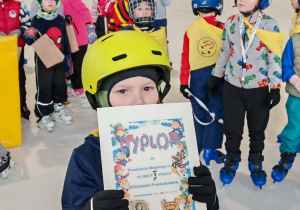 Grupa dzieci już na lodowisku cieszy się z dyplomu. Dziecko w żółtym kasku pokazuje dyplom