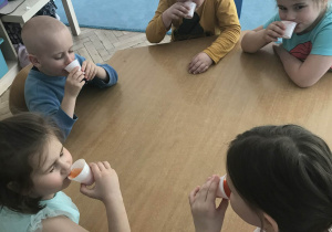 Dzieci siedzą przy stoliku i piją soki z kubeczków.