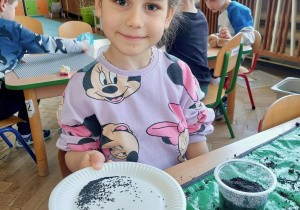 Dziecko siedzi przy stoliku i trzyma talerzyk z nasionami do wysiewu.